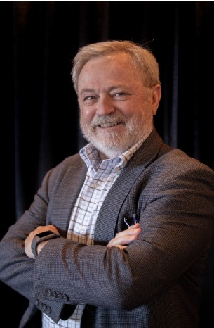 An older man with gray hair and beard stands with arms crossed, wearing a checked shirt, gray blazer, and smartwatch, in front of a dark background.