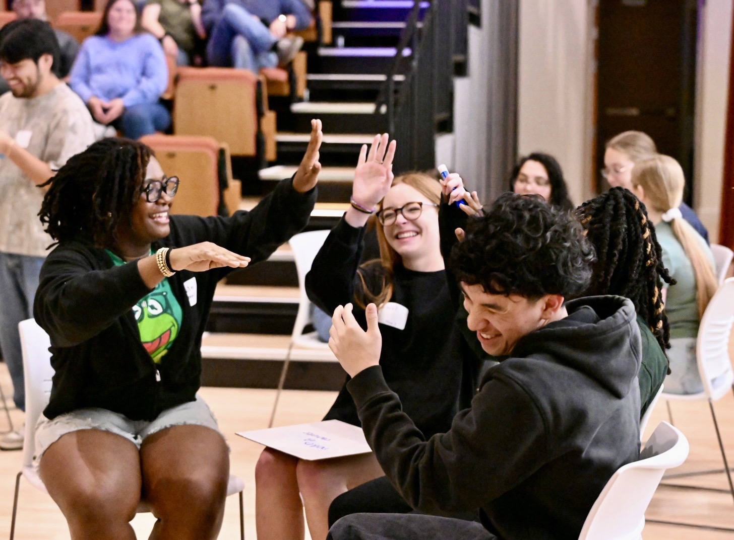 Four young adults sit in a circle indoors, smiling and giving high fives, with others seated in the background.