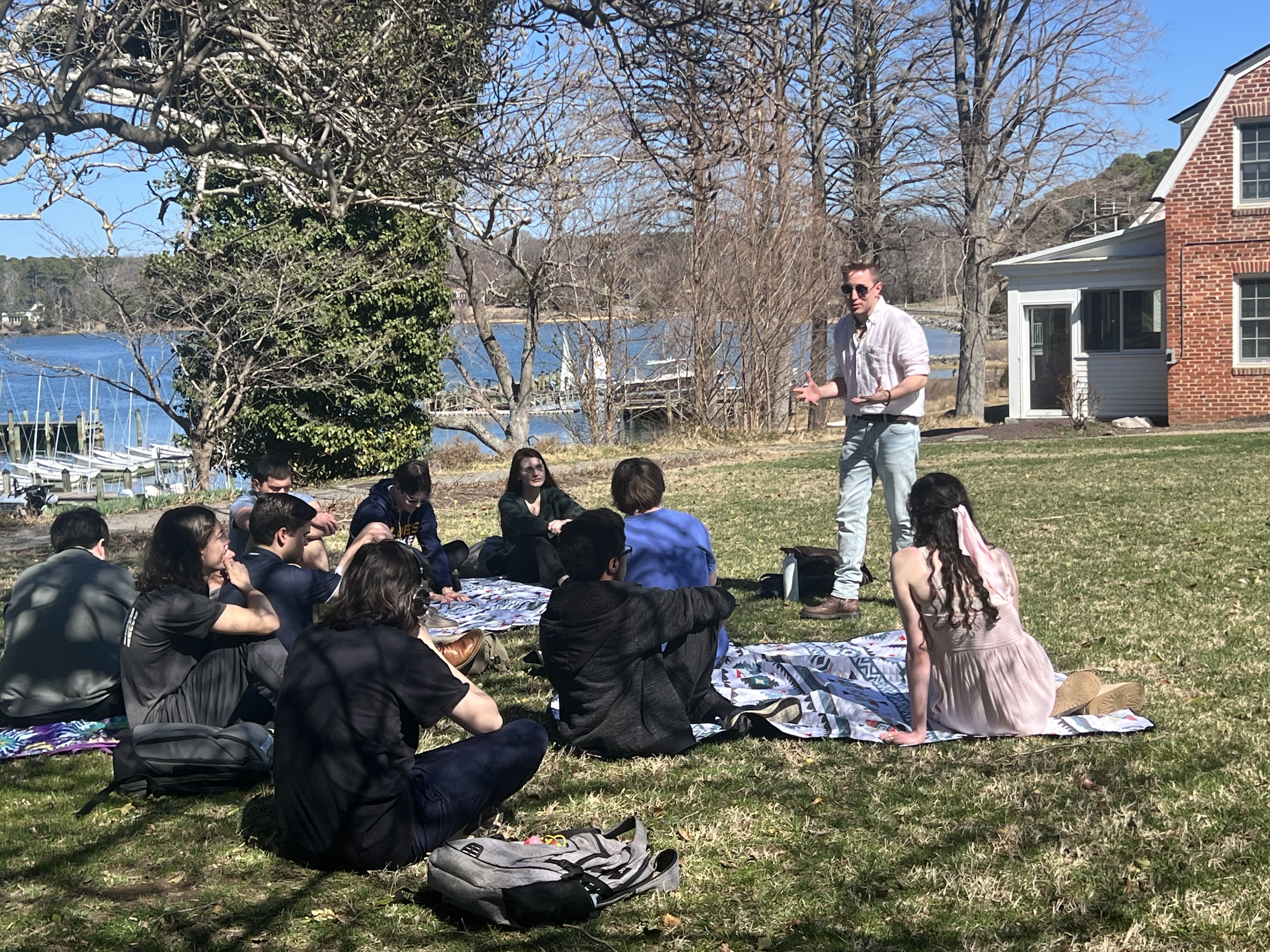 A group of people sit on blankets in the grass while a man stands and speaks to them near a lake and a brick building on a sunny day.