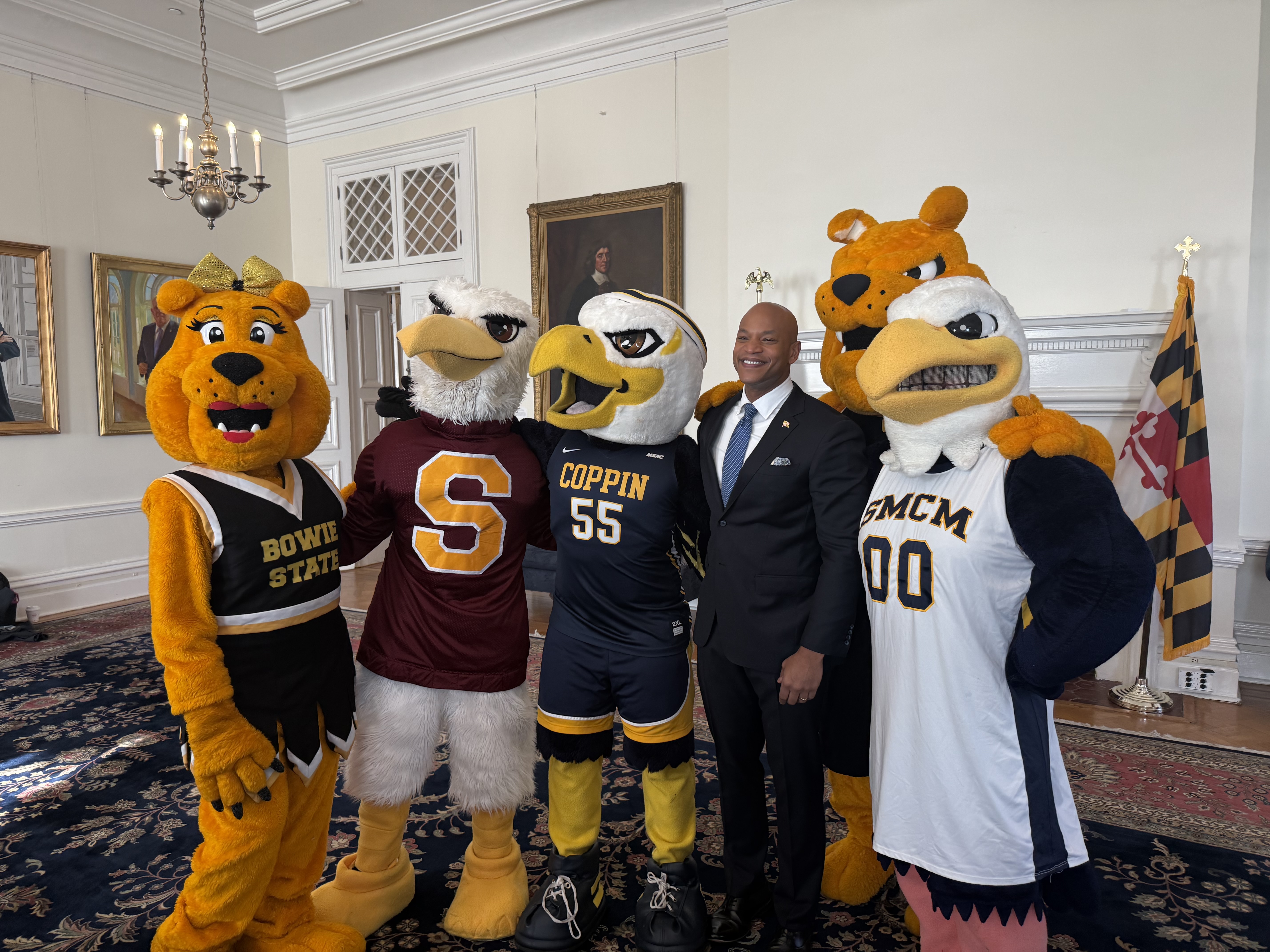 Five college mascots in uniform and a man in a suit stand together indoors, posing for a group photo. Maryland state flag is visible in the background.