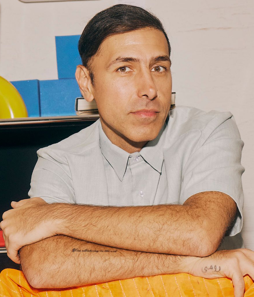A person with short dark hair and a light grey shirt sits with arms crossed on a table covered by an orange cloth, with colorful objects in the background.