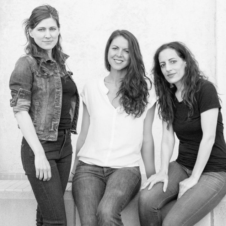 Three women pose together in casual clothing against a plain, light-colored background. Two are standing and one is seated in the center. The image is in black and white.