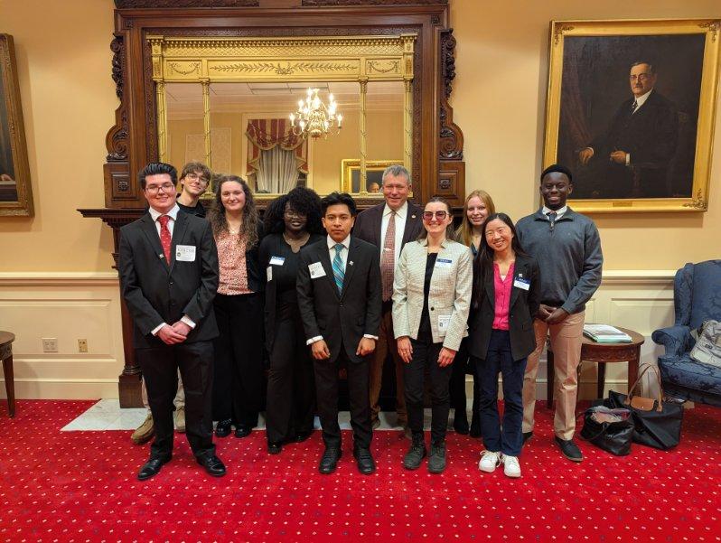 A group of eleven people in formal and semi-formal attire pose for a photo in a room with red carpet, wood paneling, a large mirror, and a portrait on the wall.