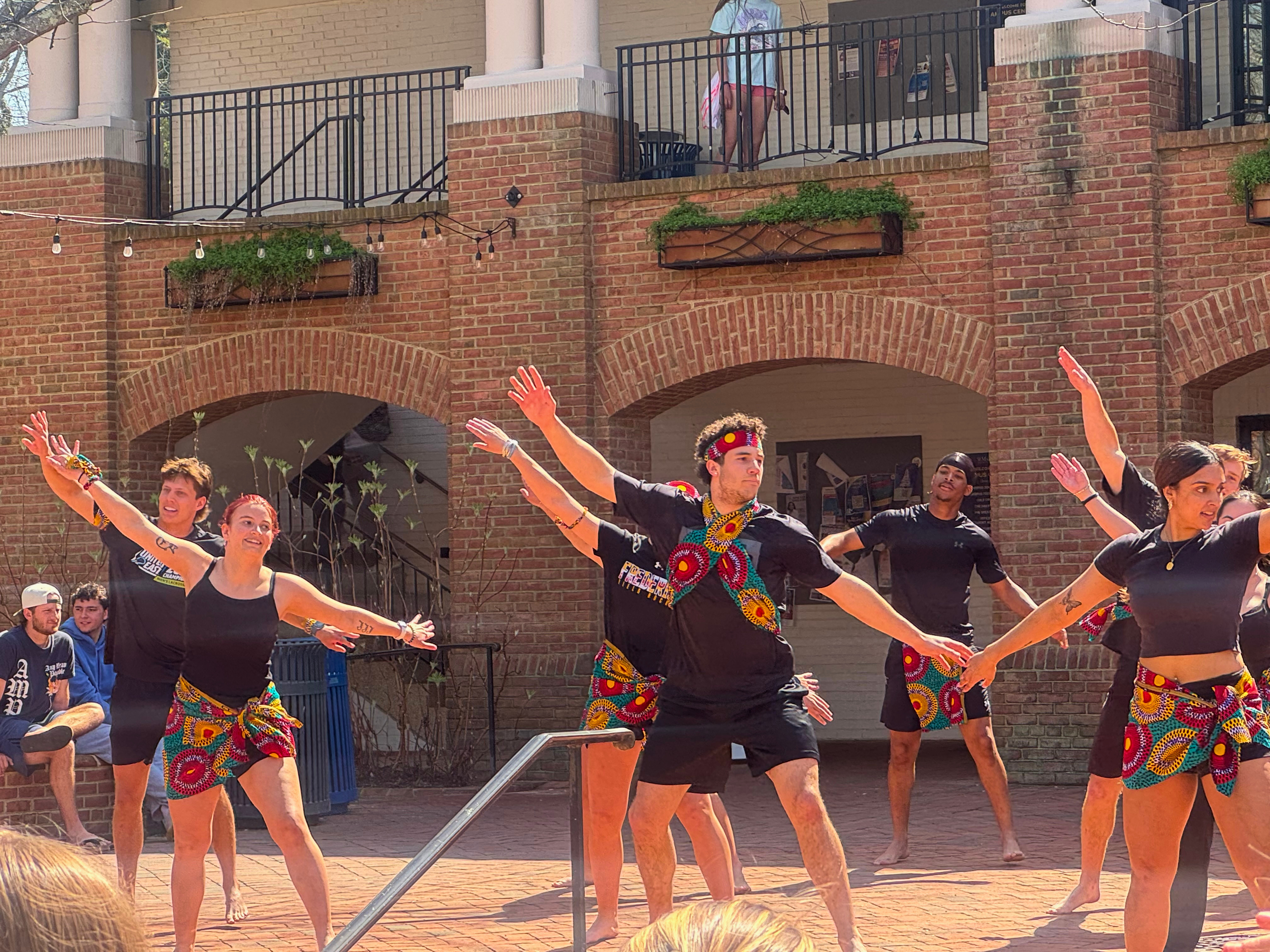 A group of dancers in black outfits with colorful patterned skirts perform outdoors in front of a brick building, arms extended in mid-movement.
