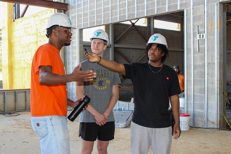 Three people wearing hard hats stand inside a building under construction; one gestures while another holds a recorder toward him.
