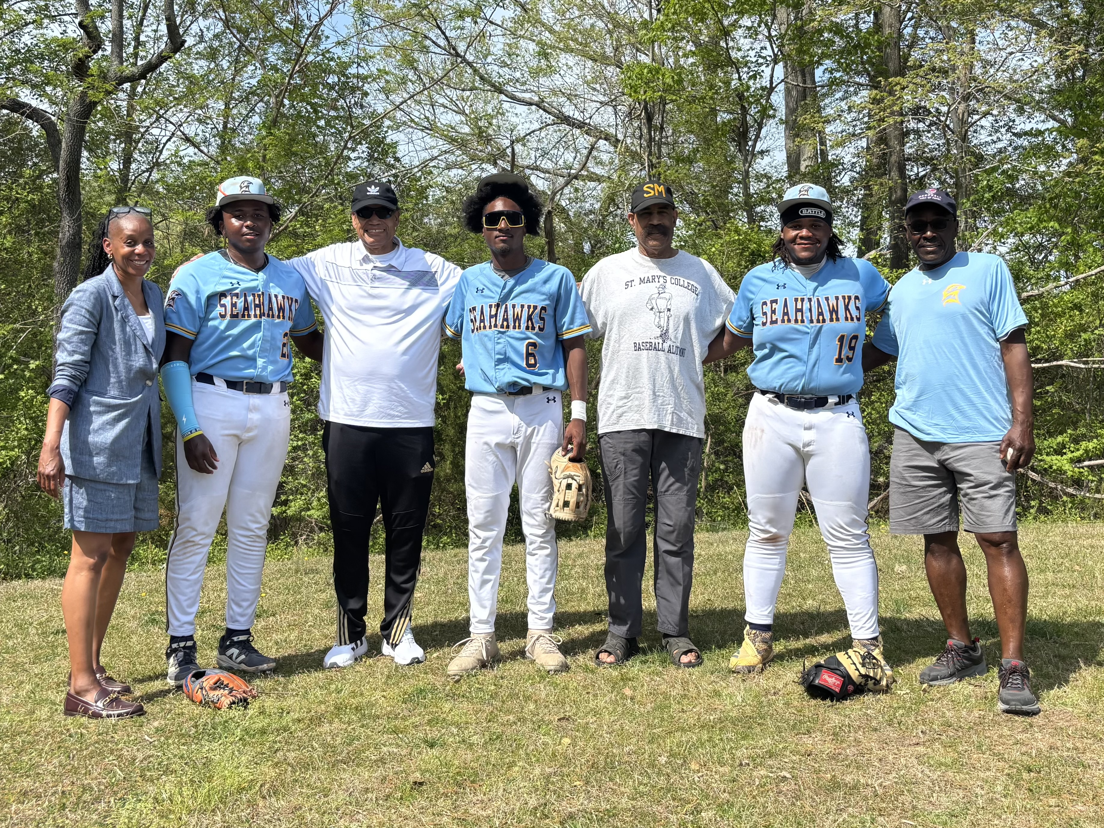 Seven people stand outdoors on grass, including three in Seahawks baseball uniforms, flanked by four adults in casual clothing. Trees and greenery are visible in the background.