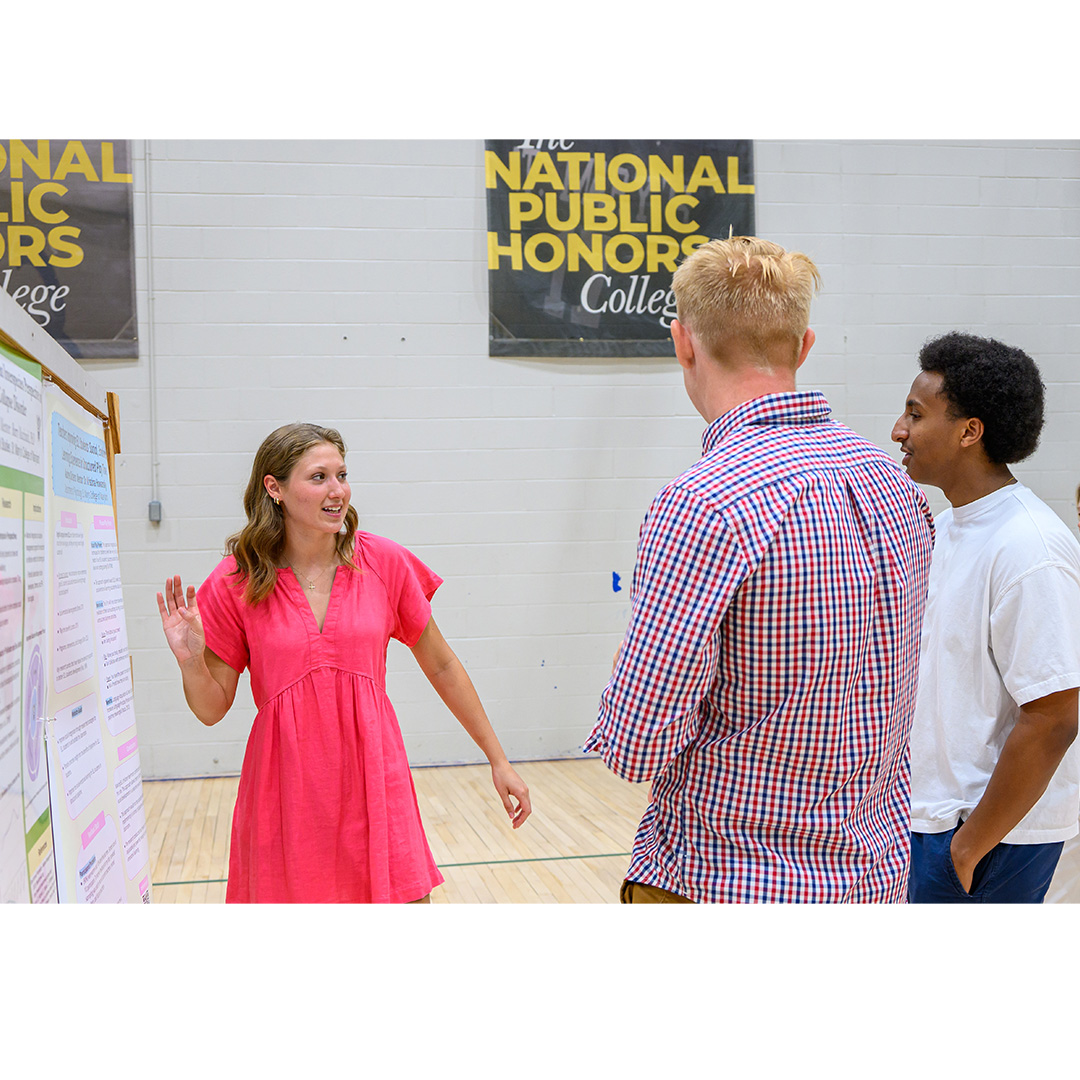 A woman in a pink dress presents a poster to two men in a gymnasium with "National Public Honors College" banners on the wall.