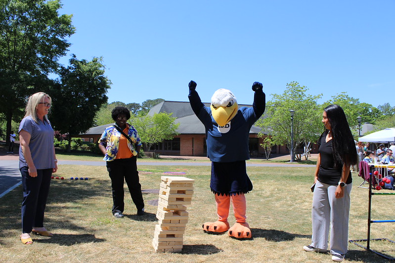 Four people, including one in an eagle mascot costume, stand around a giant Jenga tower outdoors on a sunny day. The mascot has its arms raised in celebration.