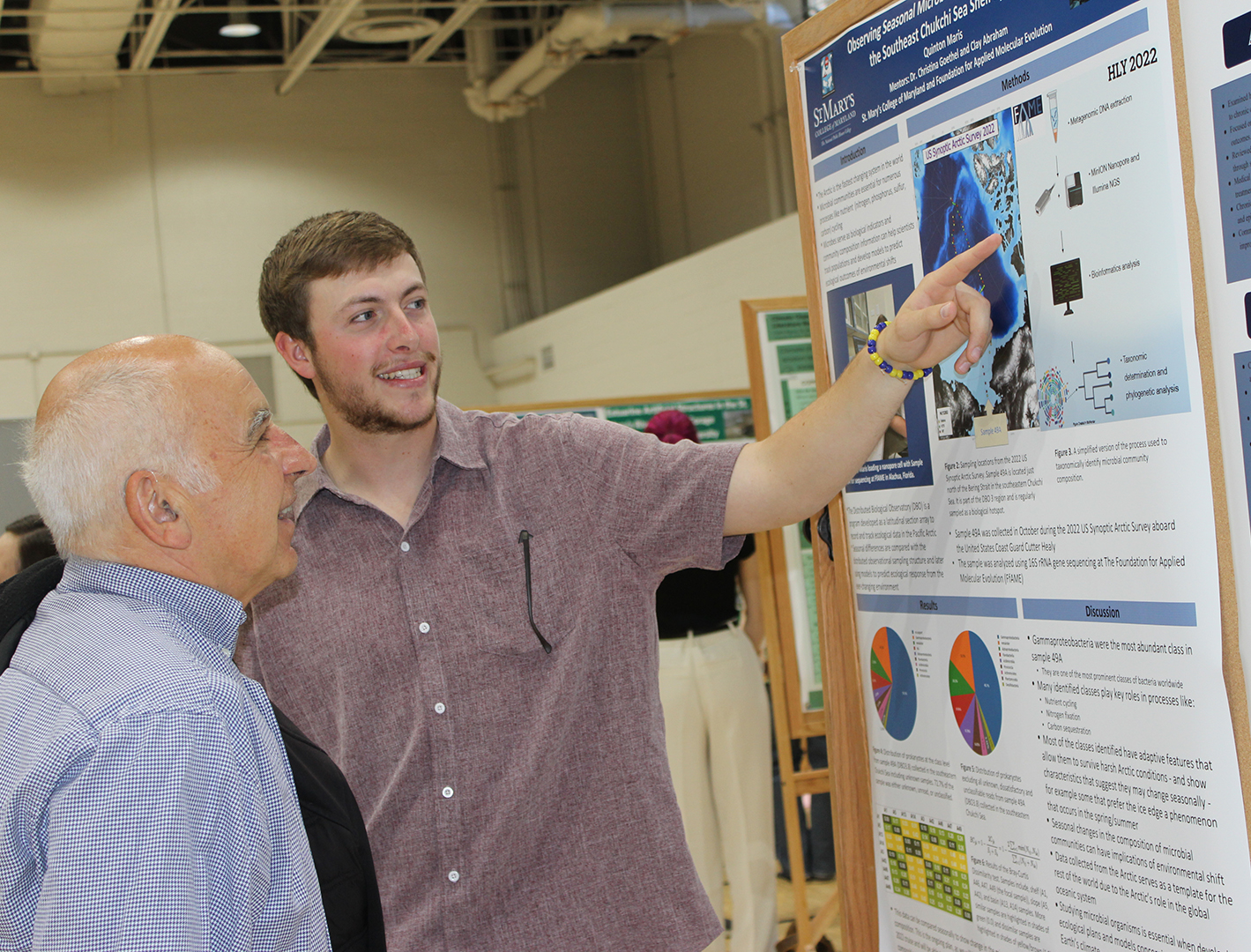 Two men stand in front of a scientific poster display; the younger man points at the poster while explaining information to the older man.
