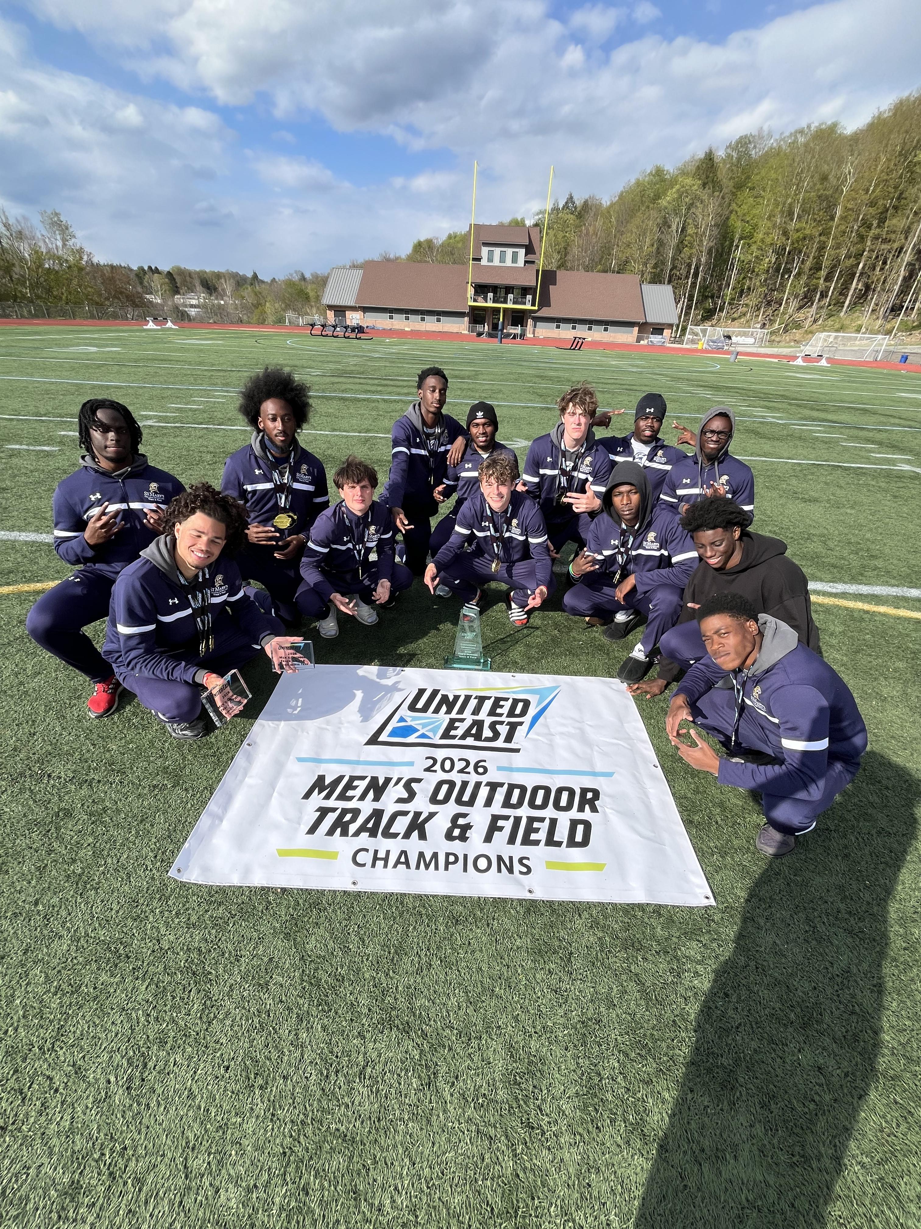 A men's track and field team poses on a field behind a banner that reads "United East 2026 Men's Outdoor Track & Field Champions."