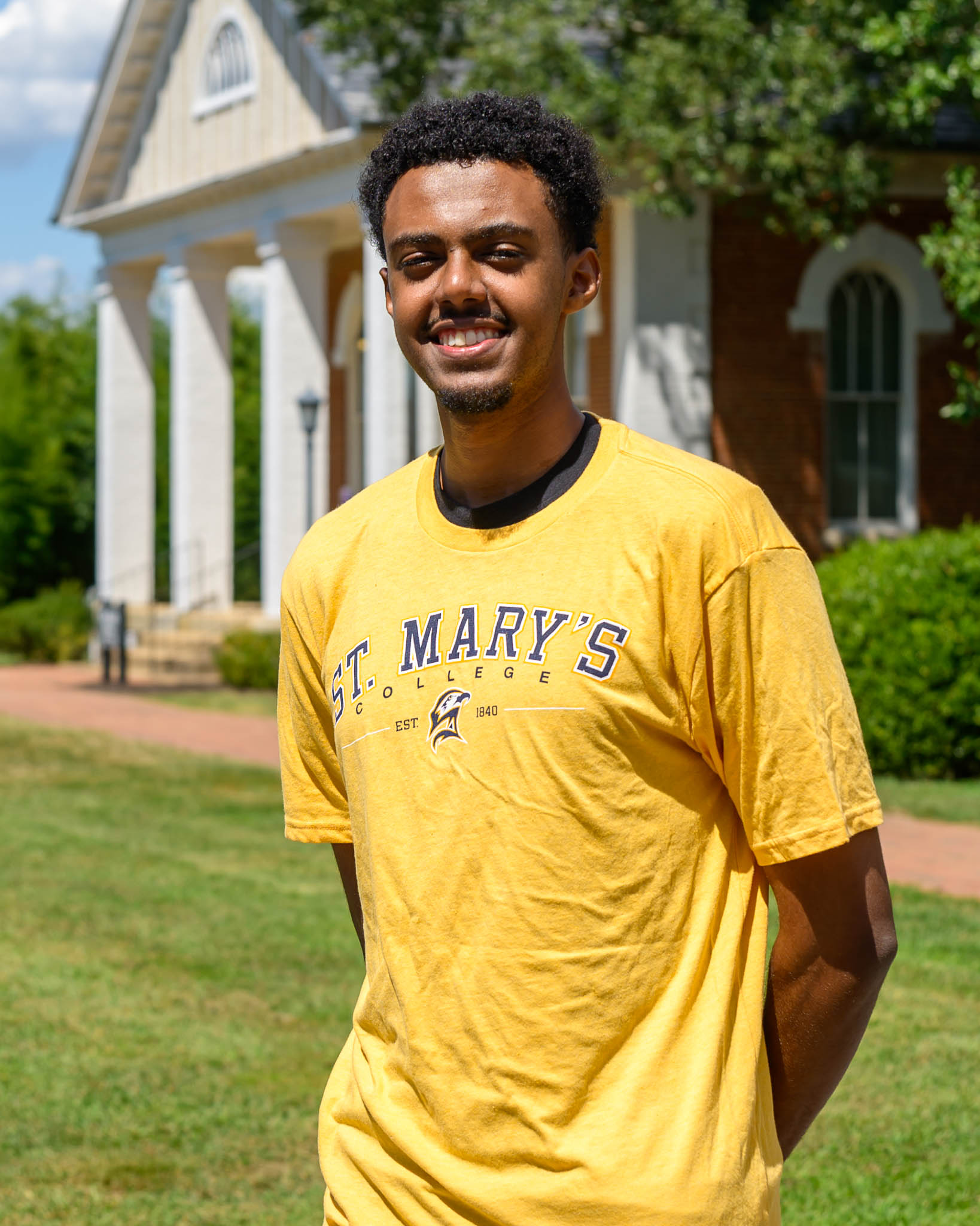 A young man in a yellow St. Mary's College T-shirt stands outside on a sunny day, smiling, with a brick building and greenery in the background.