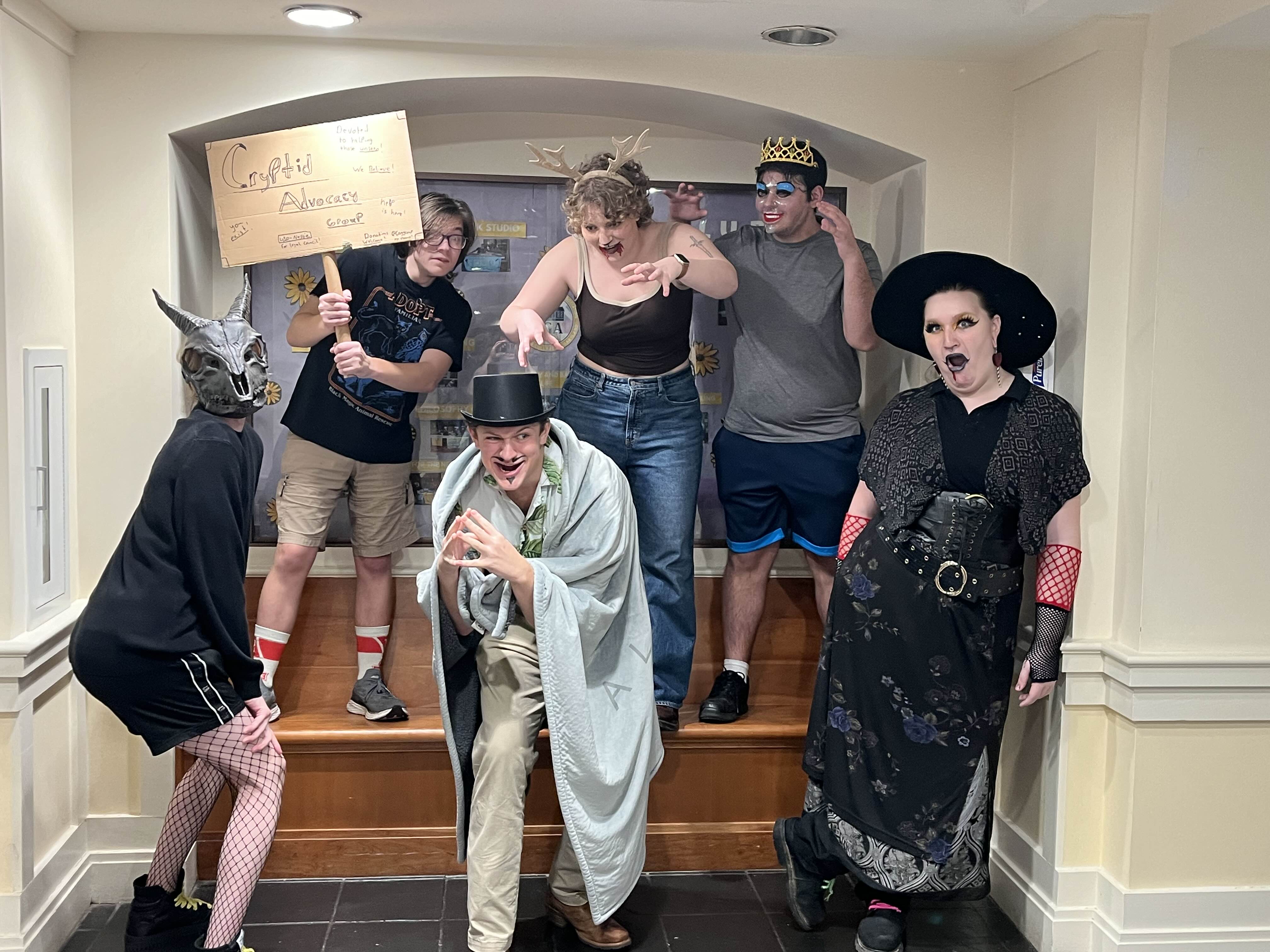 Six people in eclectic costumes pose theatrically indoors; one holds a sign reading