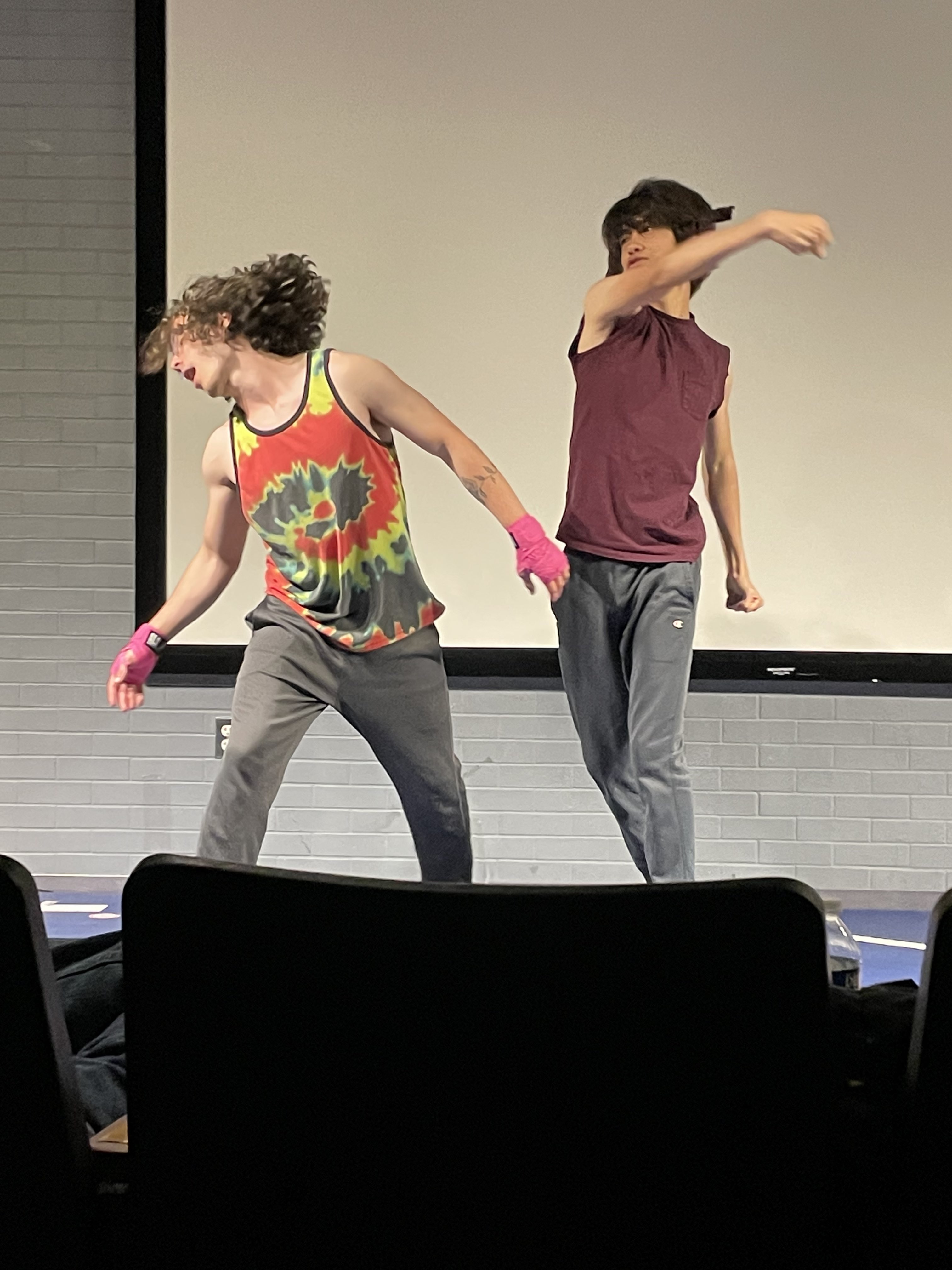 Two young people are performing on stage; one wears a tie-dye tank top and pink gloves, while the other swings an arm in a maroon sleeveless shirt.