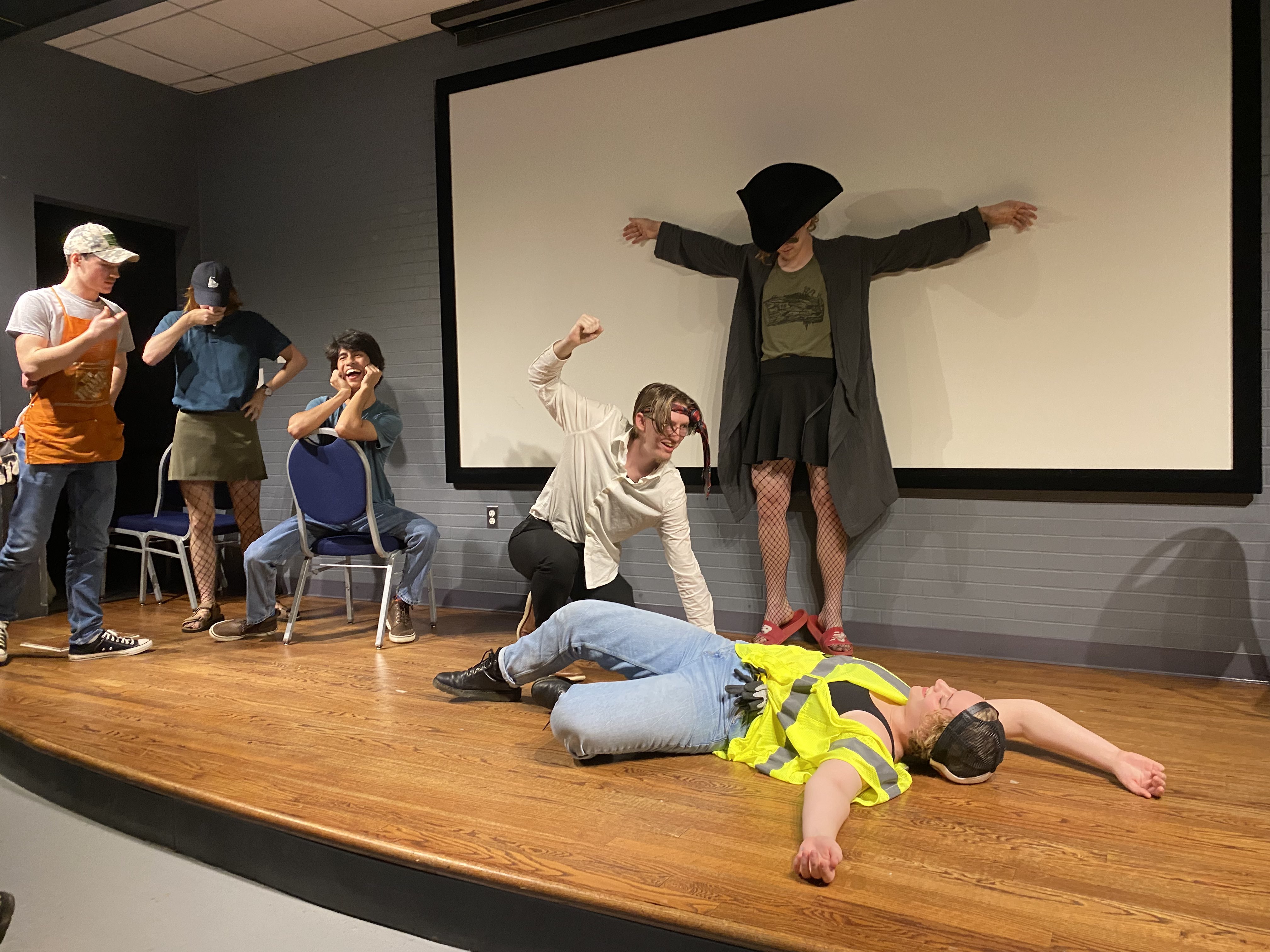 Six people perform a dramatic scene on stage; one person is on the floor in a safety vest, another raises a fist over them, while others react expressively.