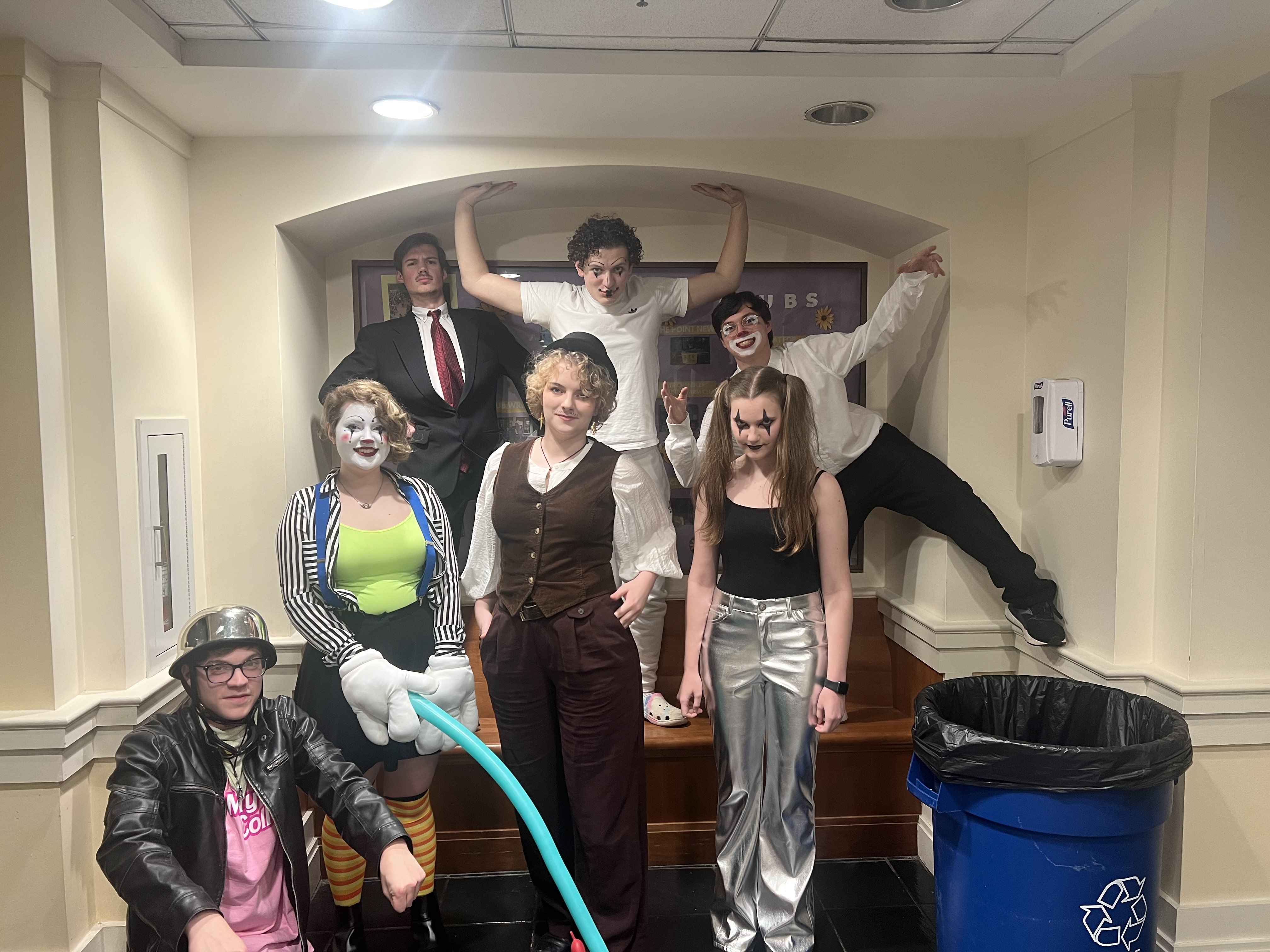 Seven people in costume pose together indoors; some wear clown makeup, hats, or shiny pants, and one person holds long balloons. They stand or sit next to a large recycling bin.
