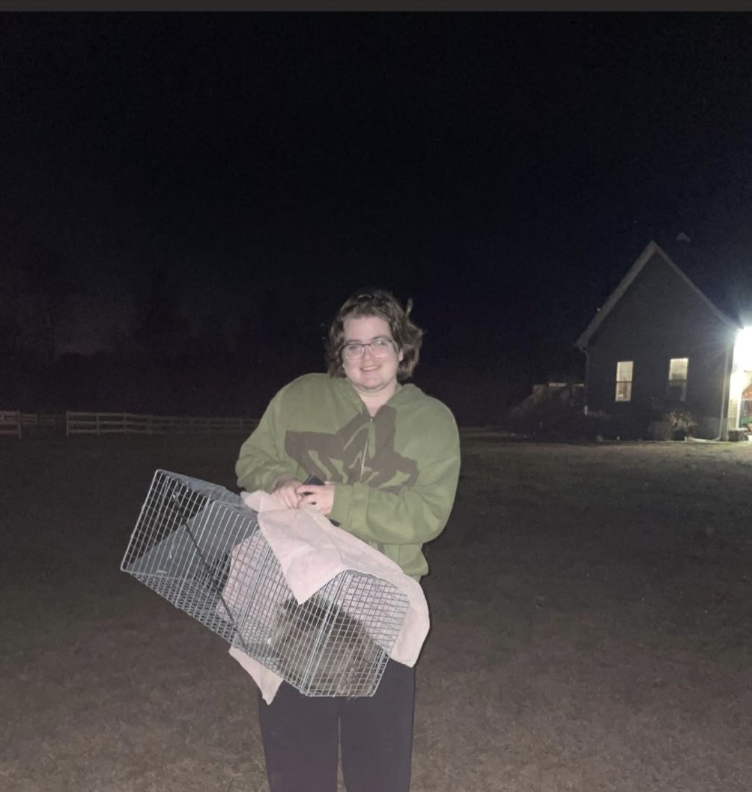 A person standing outside at night holding a metal animal trap containing an animal.