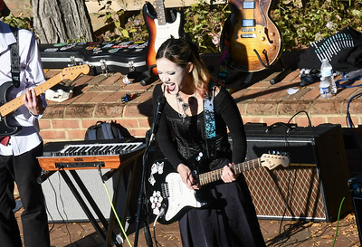 A woman plays an electric guitar and sings outdoors near a keyboard and amplifiers, with other guitars and music equipment visible in the background.