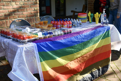 A table covered with a rainbow flag holds drinks, snacks, utensils, and hand sanitizer, set up outdoors near a brick wall.