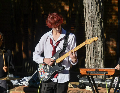 A person with red hair plays an electric guitar outdoors, wearing a white shirt and red tie. A keyboard and drum set are also visible.