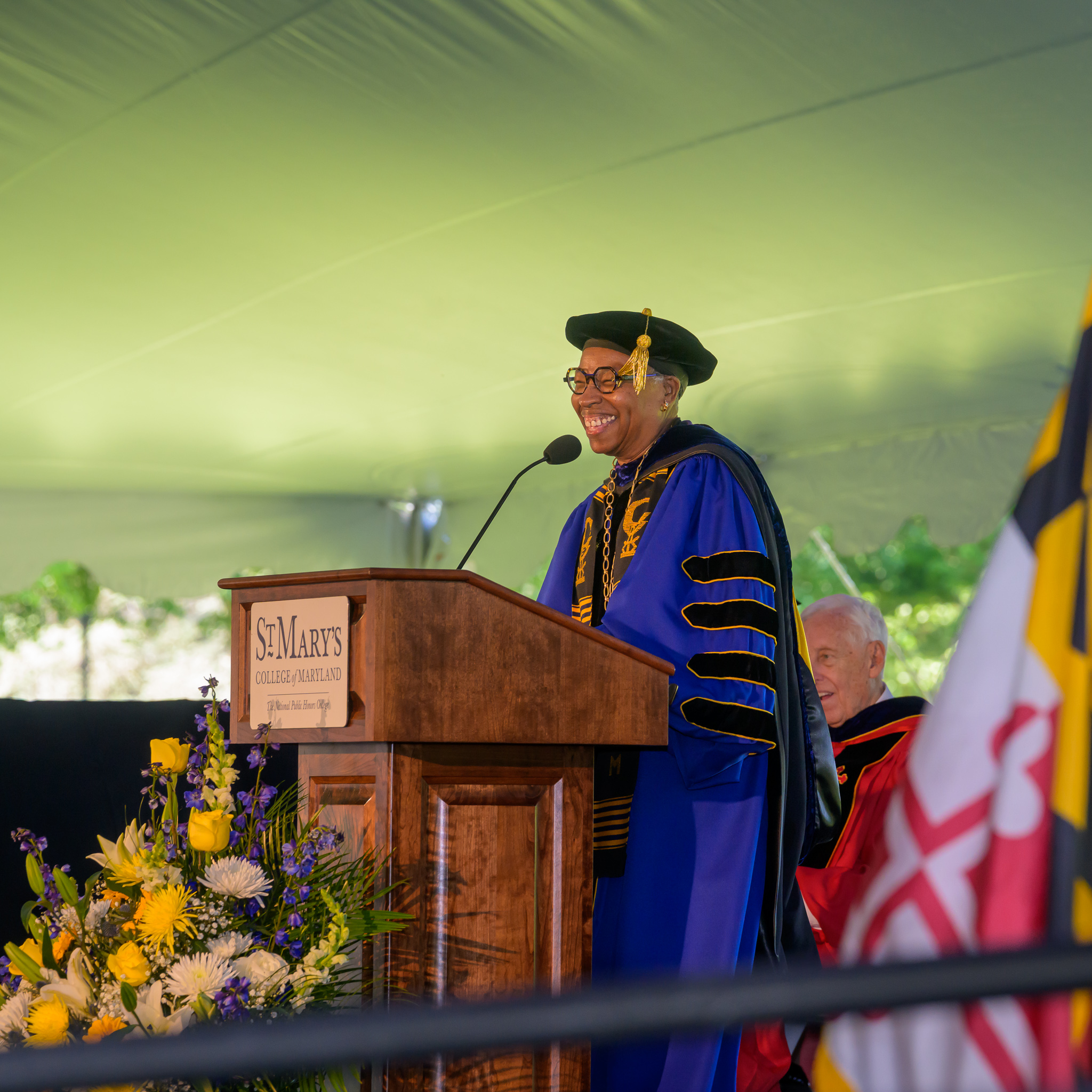 A person in academic regalia speaks at a podium labeled "St. Mary's College of Maryland" during a graduation ceremony, with flowers and the Maryland flag nearby.