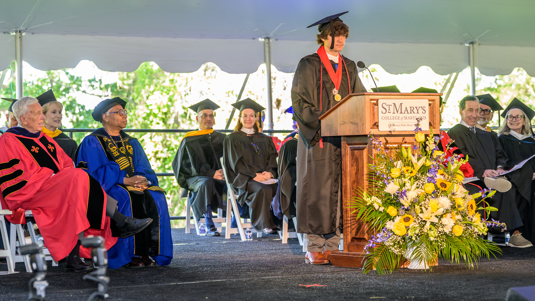 A graduate in cap and gown speaks at a podium labeled "St. Mary's College," while faculty members in academic regalia sit on stage under a tent.
