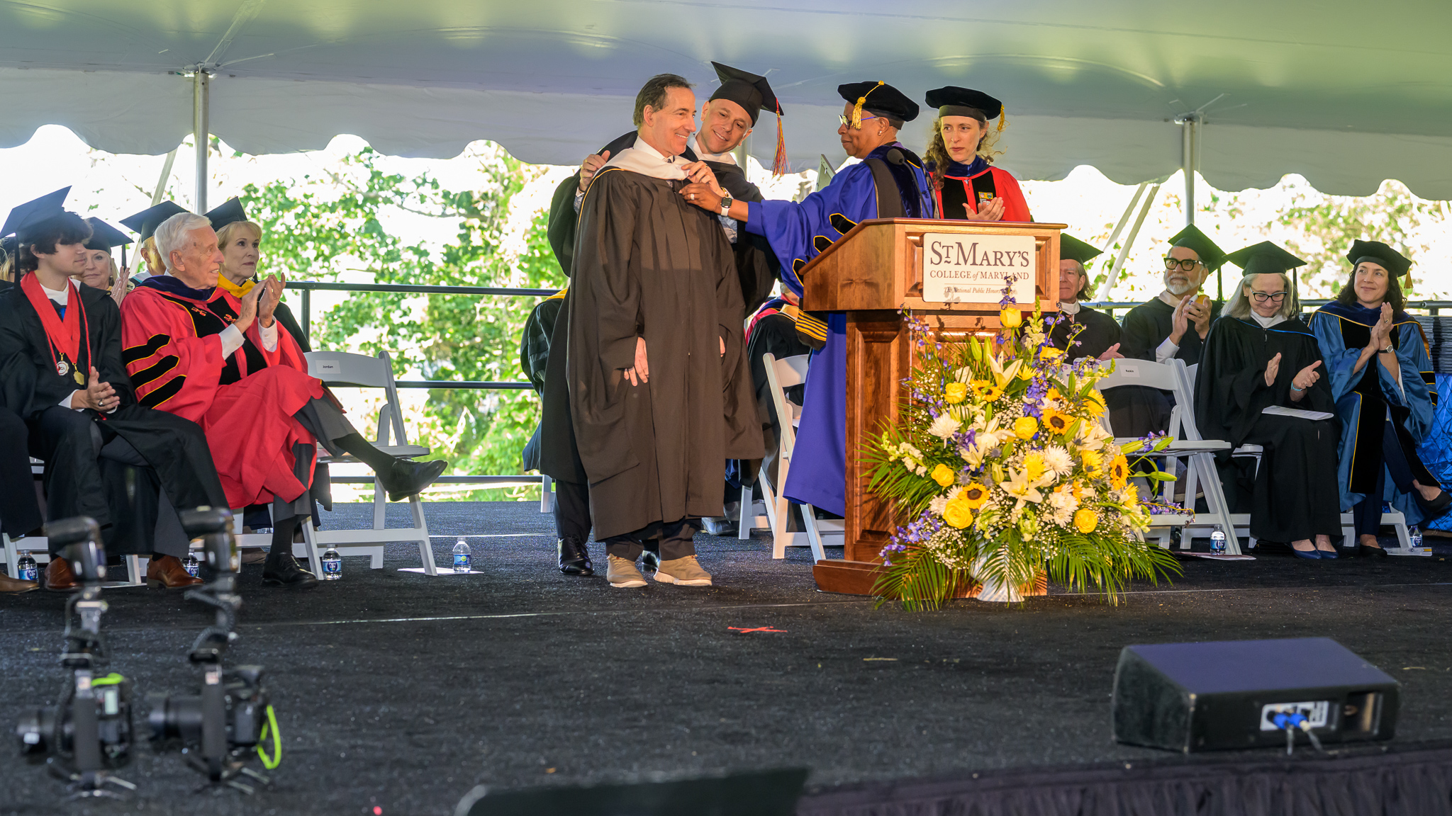 A speaker in a gown is being hooded by faculty on stage during a St. Mary's College graduation ceremony, with other faculty and attendees seated nearby.