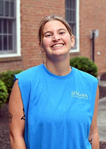 A smiling person wearing a blue sleeveless "St. Mary's College of Maryland" shirt stands outside near a brick building and shrubs.