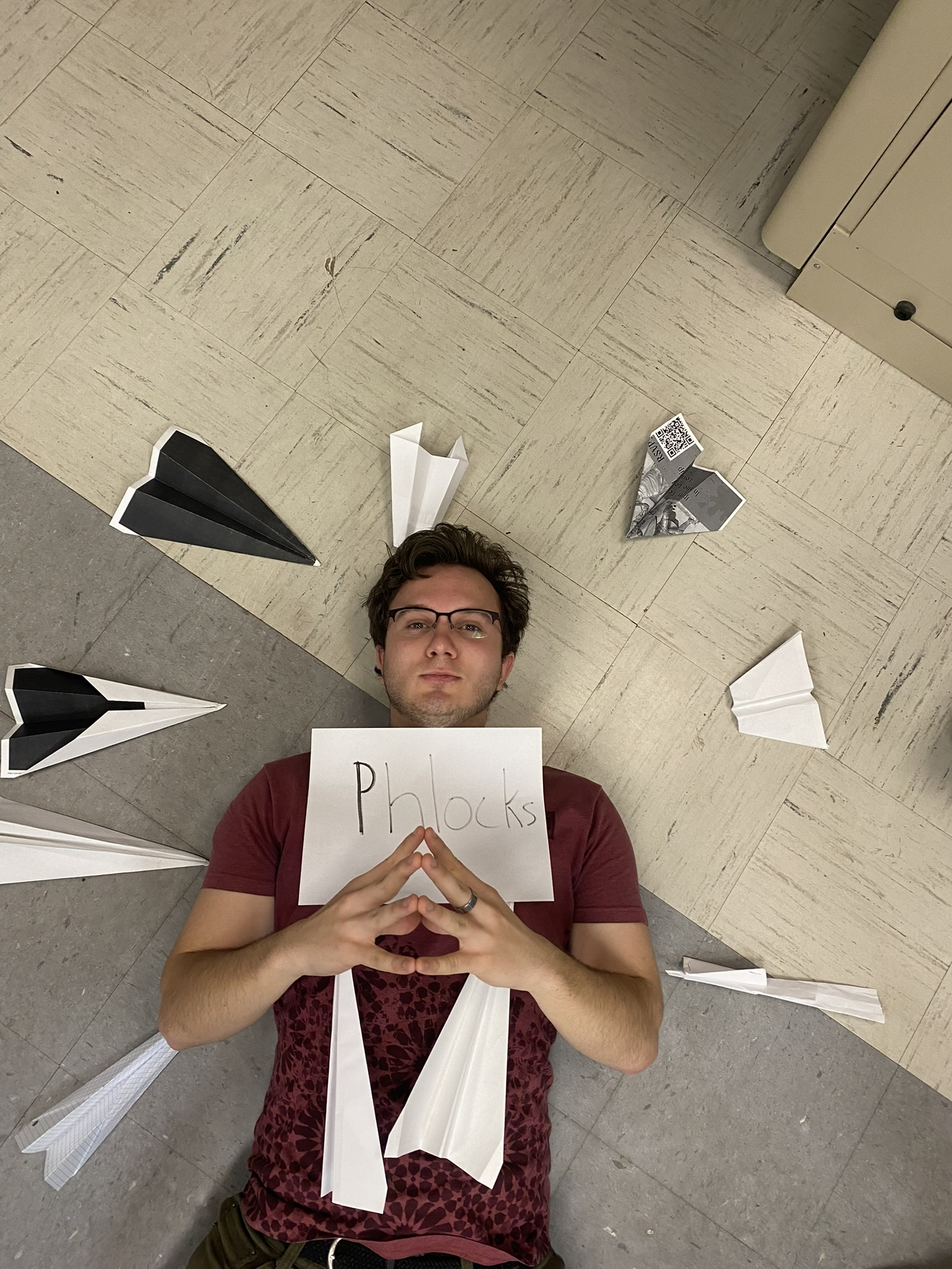 A person lies on a tiled floor surrounded by paper airplanes, holding a sign that reads
