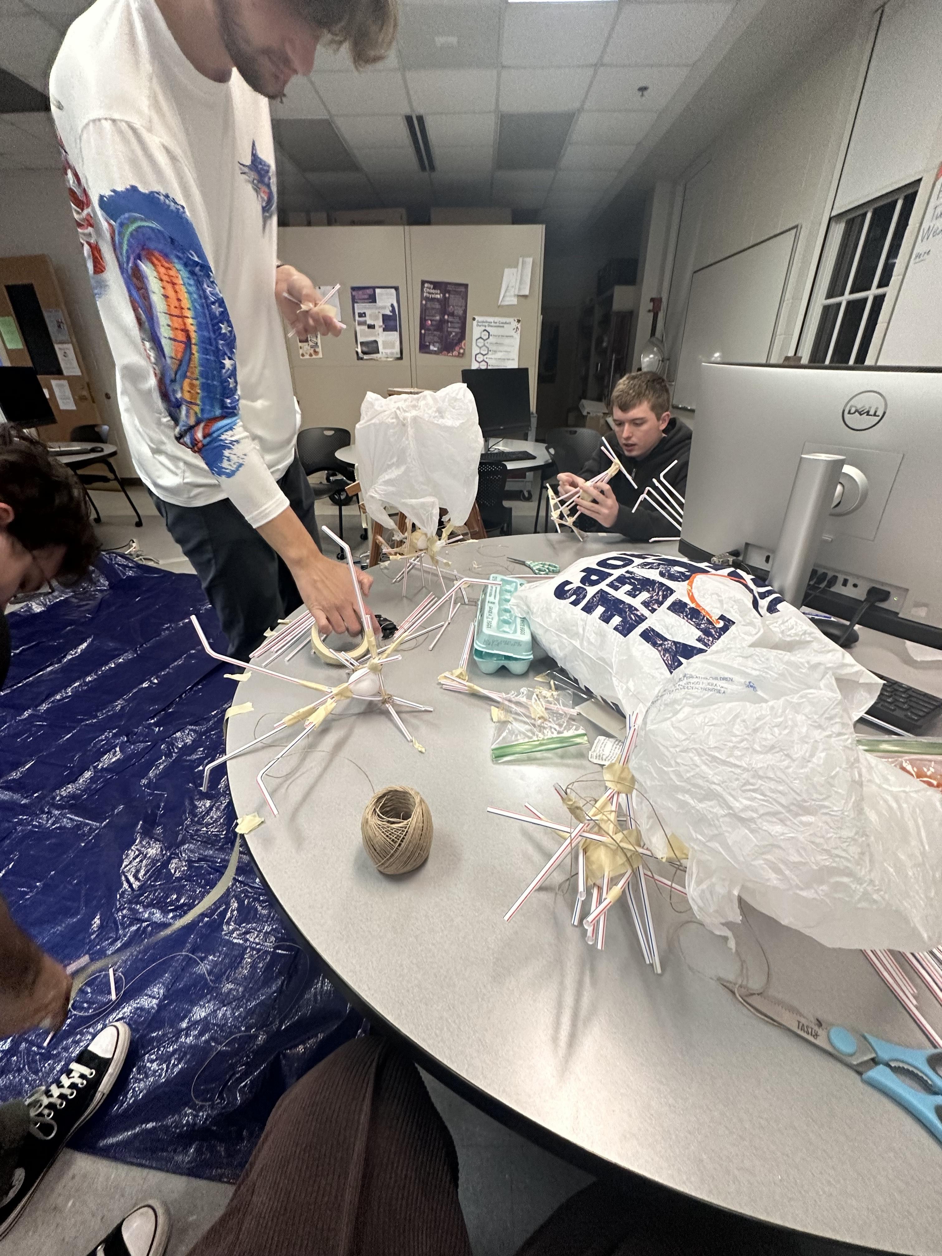 Three people work on a project using straws, string, and plastic bags at a round table in a classroom setting, with computers and materials visible.