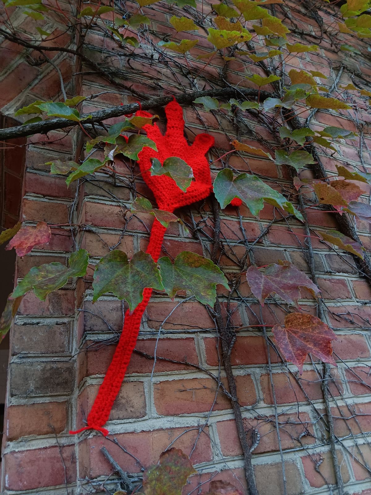 A crocheted red hand is attached to a brick wall covered with green and brown ivy leaves.