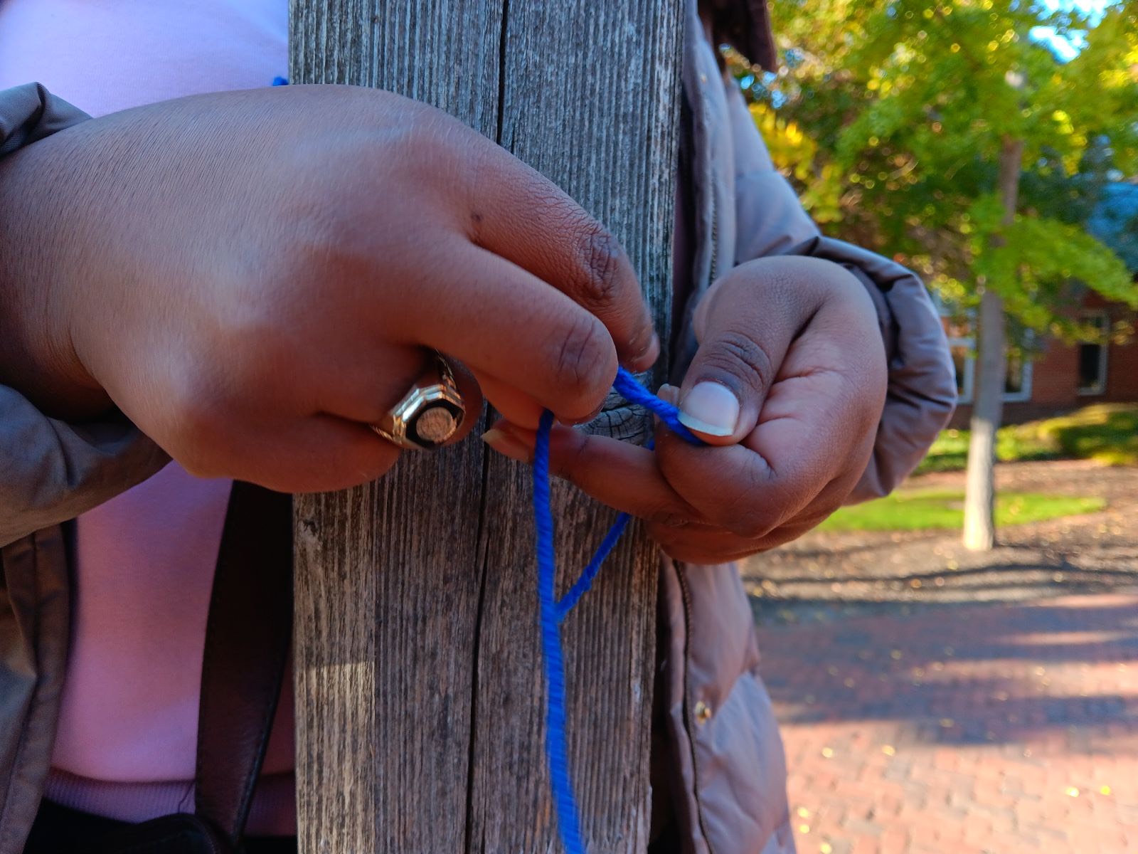 A person’s hands tying blue yarn around a weathered wooden post outdoors, with some trees and brick pavement visible in the background.