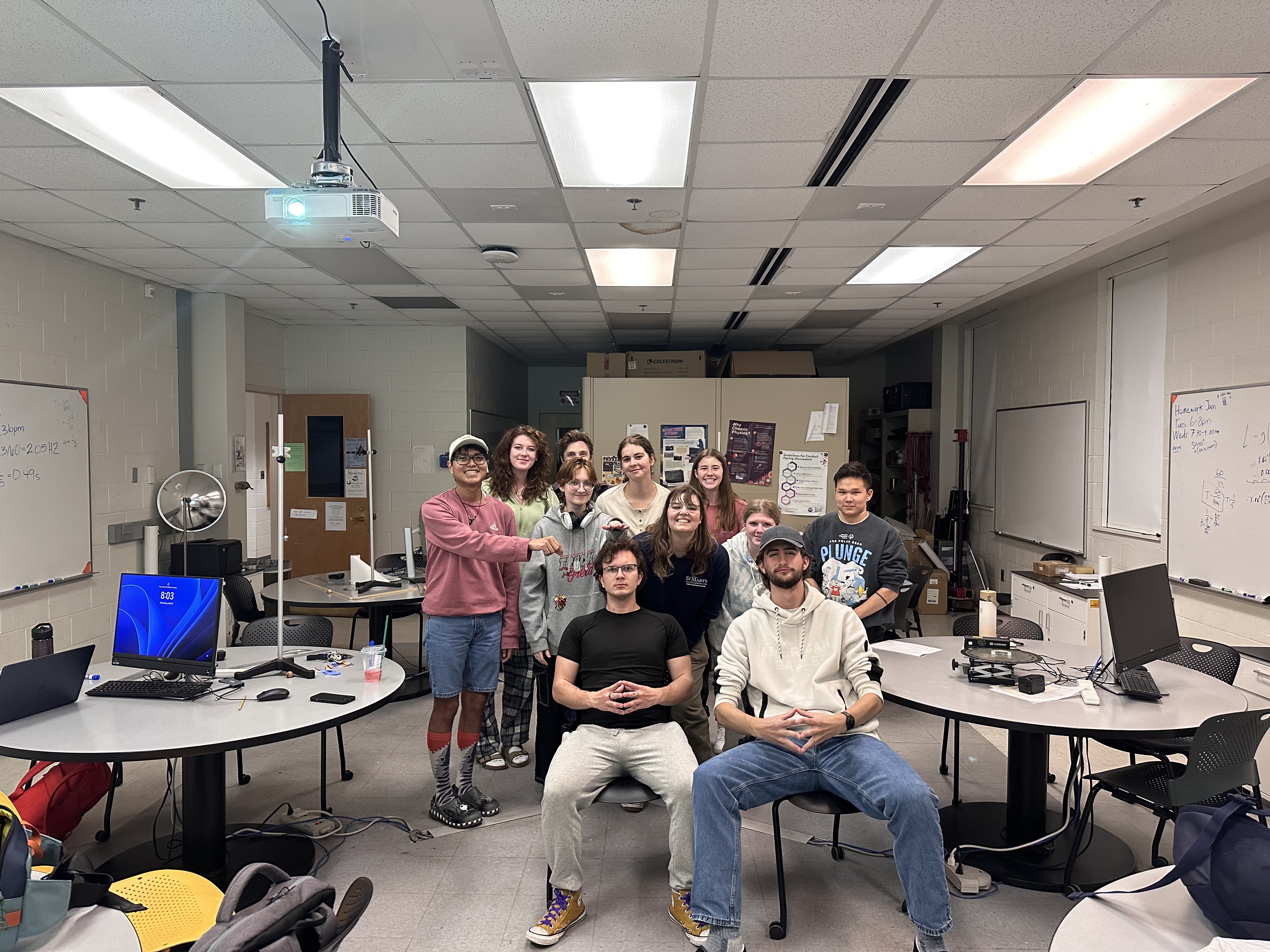A group of twelve people pose for a photo in a classroom with computers, desks, and whiteboards visible in the background.