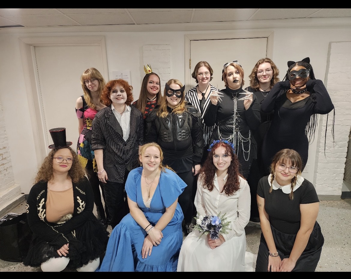 A group of thirteen people pose indoors wearing various costumes, including black outfits, face paint, cat ears, a top hat, a blue dress, and a white gown holding flowers.
