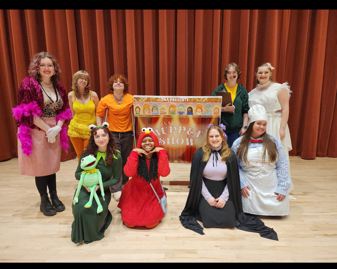 A group of people in Muppet character costumes pose in front of a puppet show sign with a red curtain backdrop.