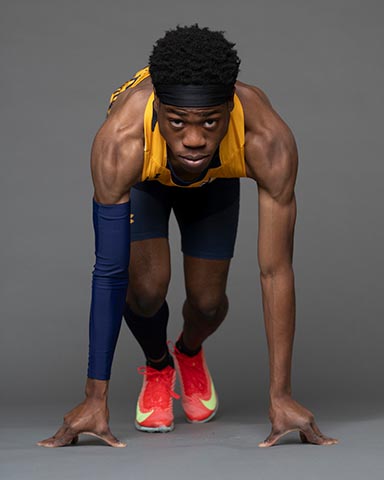 A male athlete in a gold jersey and blue shorts is in a sprinter’s starting position, looking forward against a neutral background.