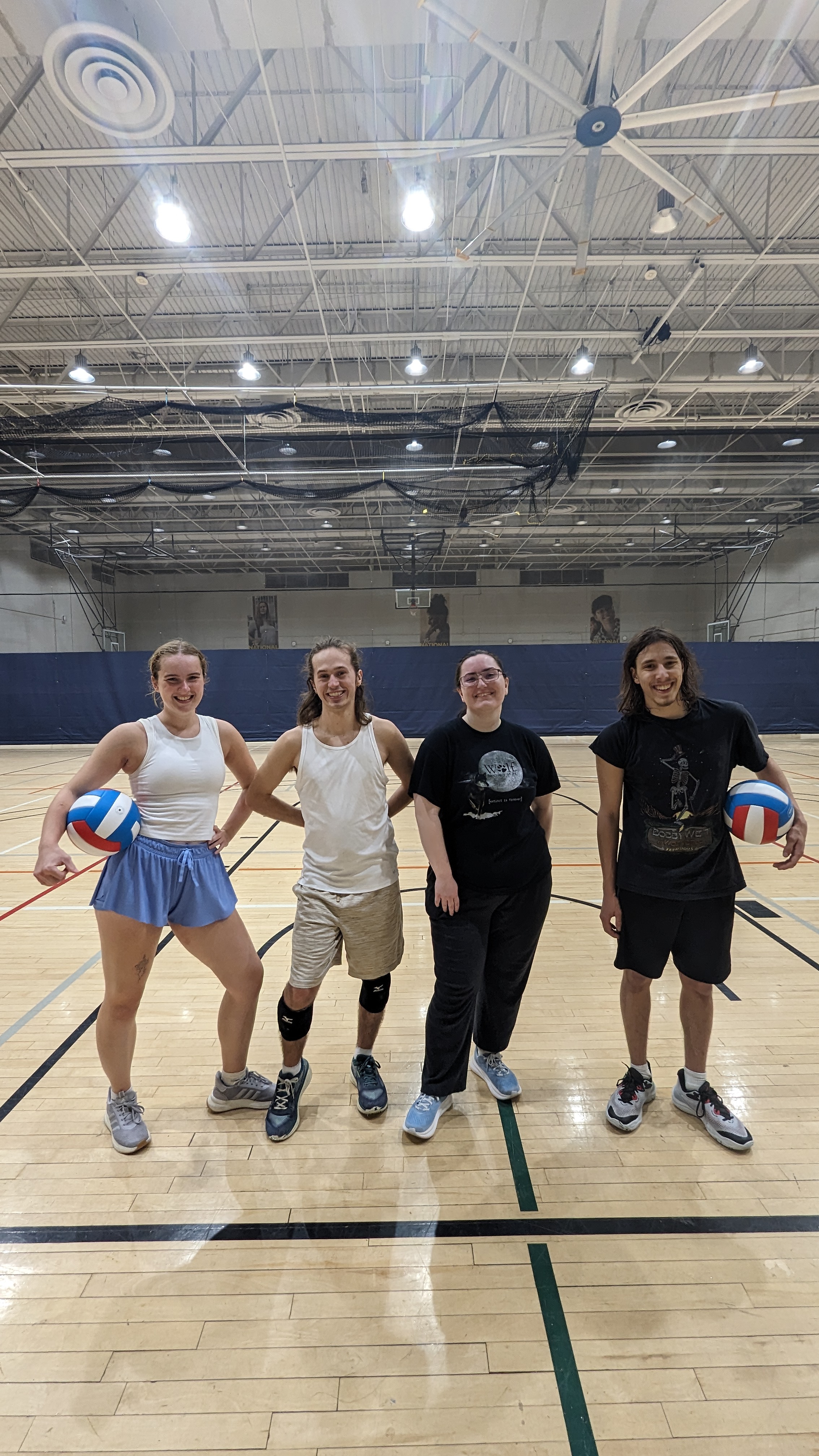 Four people standing on an indoor gym court holding volleyballs, posing and smiling for the camera.