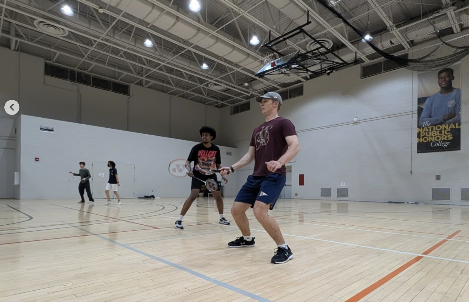 Two men play doubles badminton in a gym while two others play in the background. The gym has high ceilings, lights, and a large wall banner.