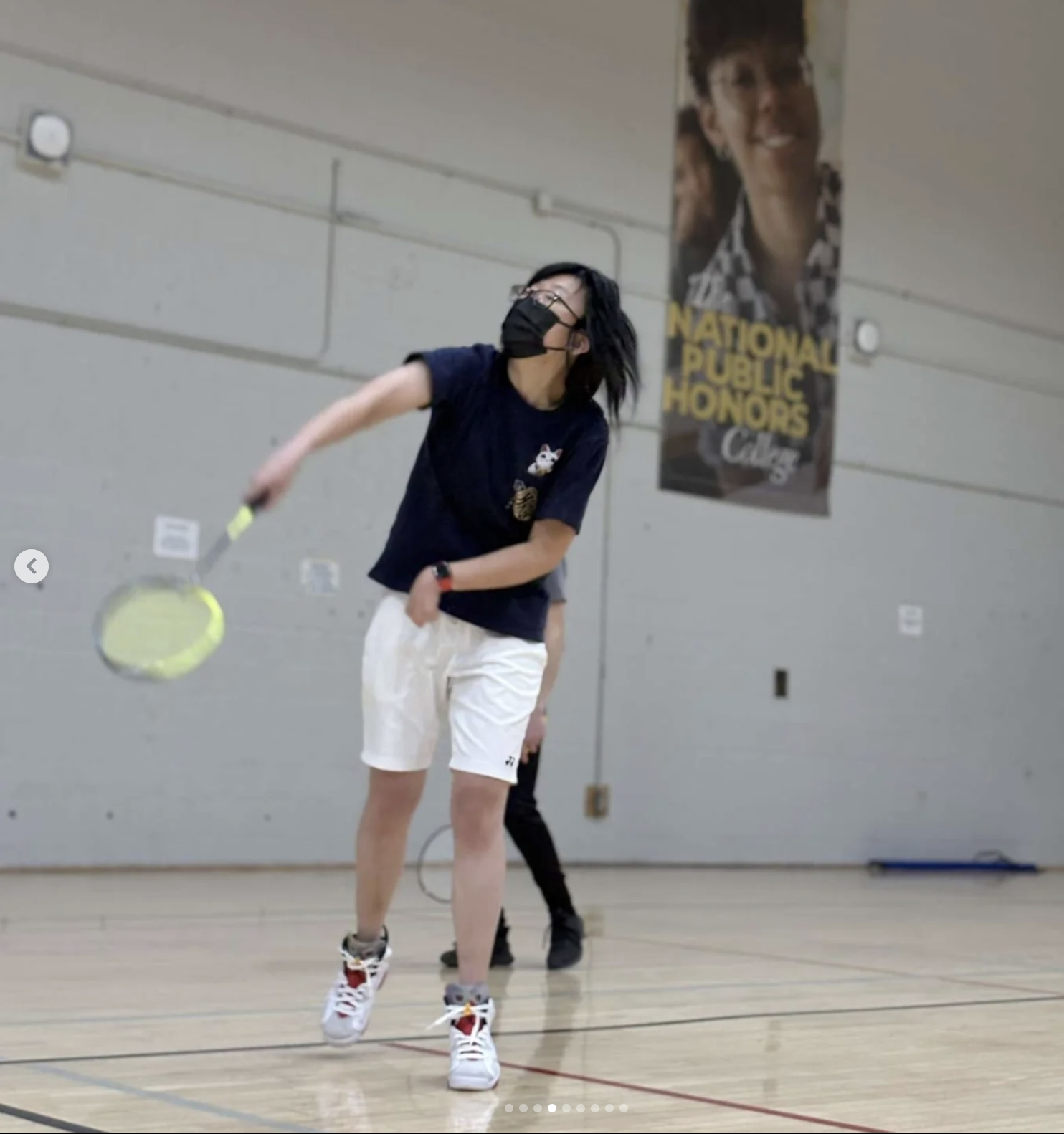 A person wearing a mask, navy shirt, and white shorts swings a badminton racket in a gymnasium. A banner hangs on the wall in the background.