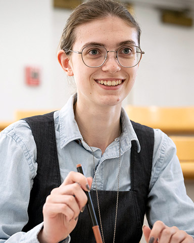 A person with glasses and long hair pulled back is smiling while holding a pencil, seated in a classroom with yellow benches in the background.