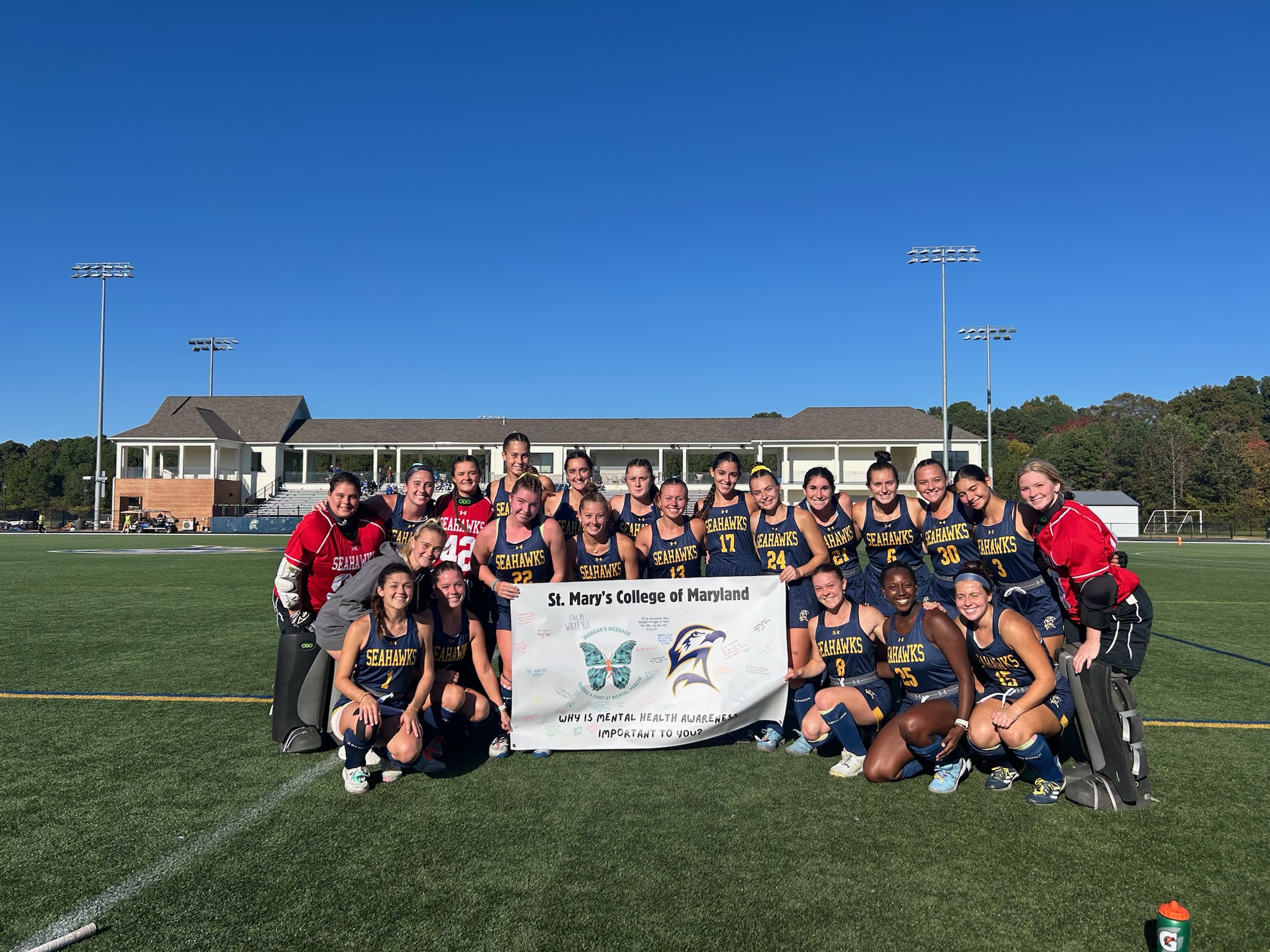 A women's field hockey team poses on an outdoor field holding a St. Mary's College of Maryland banner, with two goalies in full gear at each end.