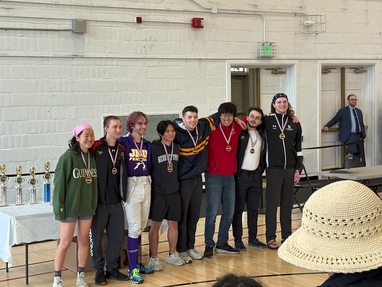 Eight people wearing medals pose for a group photo indoors near a table with trophies; a person in a hat is visible in the foreground.