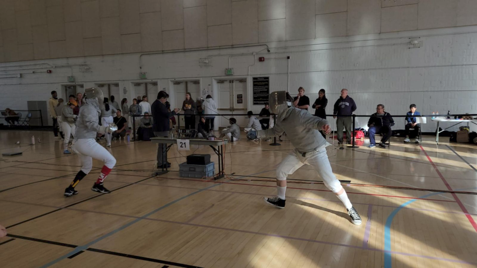 Two fencers in protective gear compete in a gymnasium as referees and spectators observe from the sidelines.