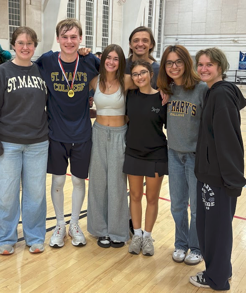 Seven young people stand together in a gym, some wearing St. Mary’s attire. One person has a medal. They are smiling and posing for a group photo.