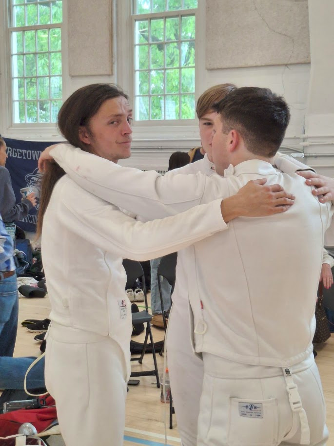 Three fencers in white suits embrace in a gym setting, with windows and chairs visible in the background.