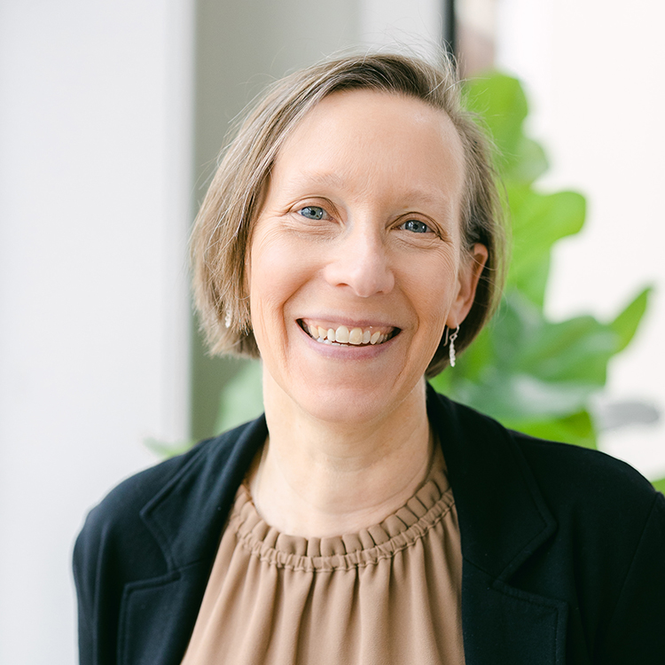 Professor Aileen Bailey, smiling female with short blond hair and blue eyes, wearing a brown shirt and black jacket in front of a blurred background of white with some greenery