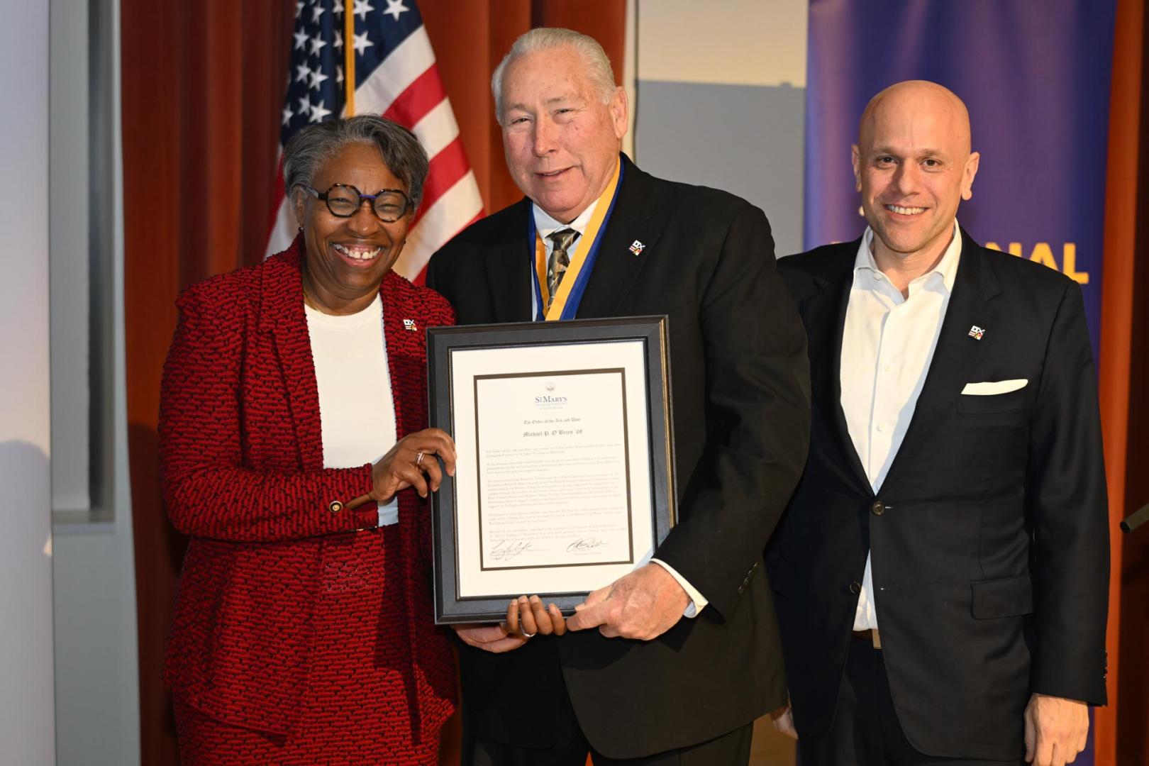 Three people stand together, smiling. The woman on the left is holding a framed document with the man in the center. An American flag and a blue banner are in the background. The man on the right is wearing a suit and has a pin on his lapel.