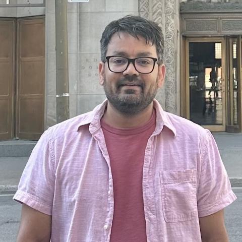 Man wearing glasses and a light pink shirt stands in front of an ornate building entrance.