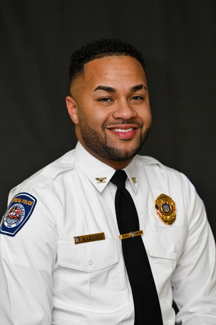 A man in a white police uniform with badges and a name tag that reads "Martinez" smiles against a dark backdrop.