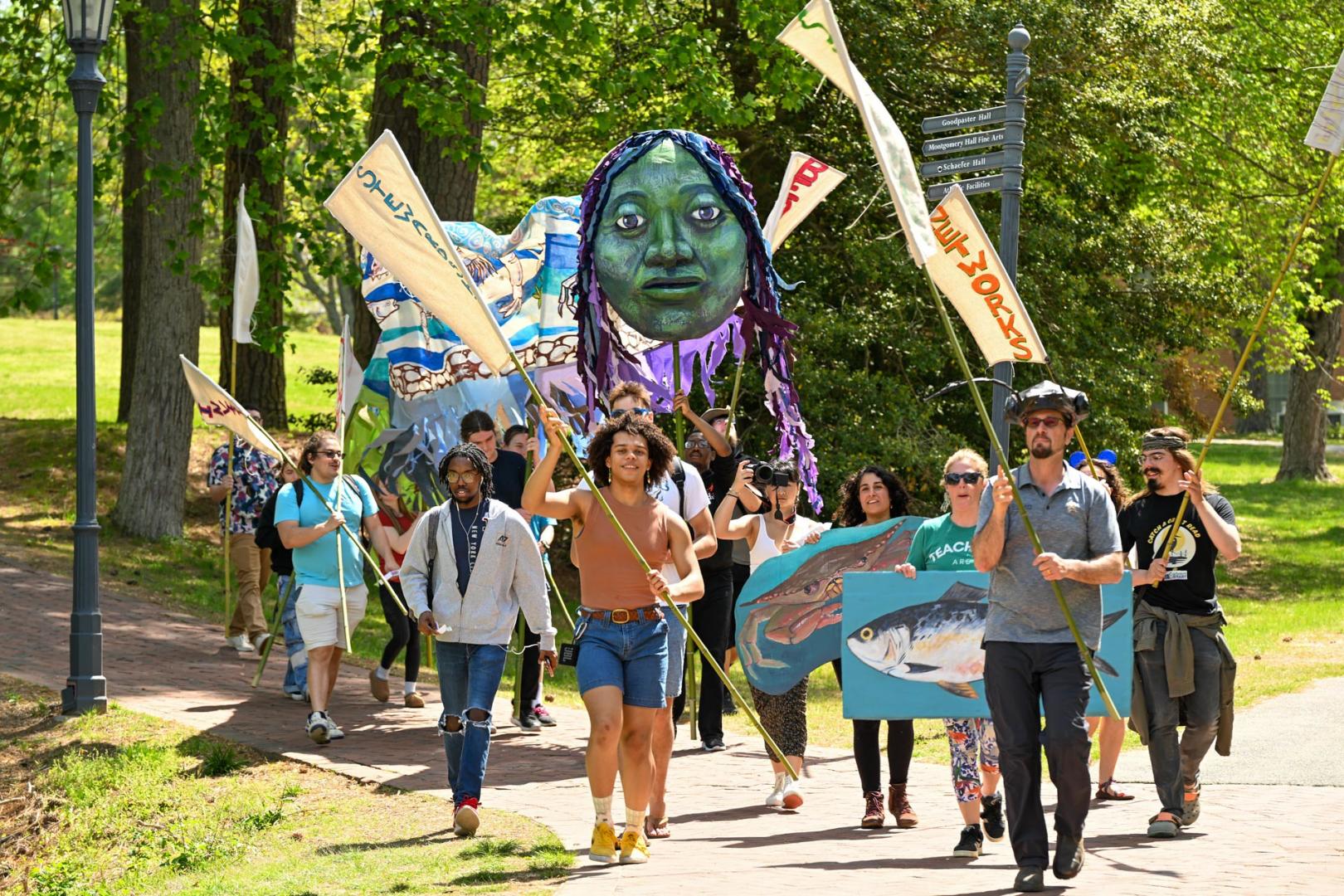 A group of people march outdoors holding banners, flags, a large puppet head, and a cardboard fish, with trees and greenery in the background.