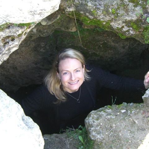 A woman with blonde hair and a black top stands inside a rocky hole, looking up and smiling at the camera. Moss and rocks surround the opening.